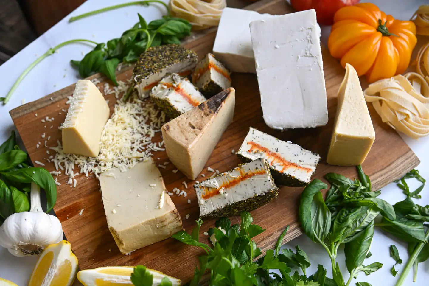 Eight assorted dairy-free cheeses on a wooden cutting board with fresh basil and heirloom tomatoes along the edge of the board