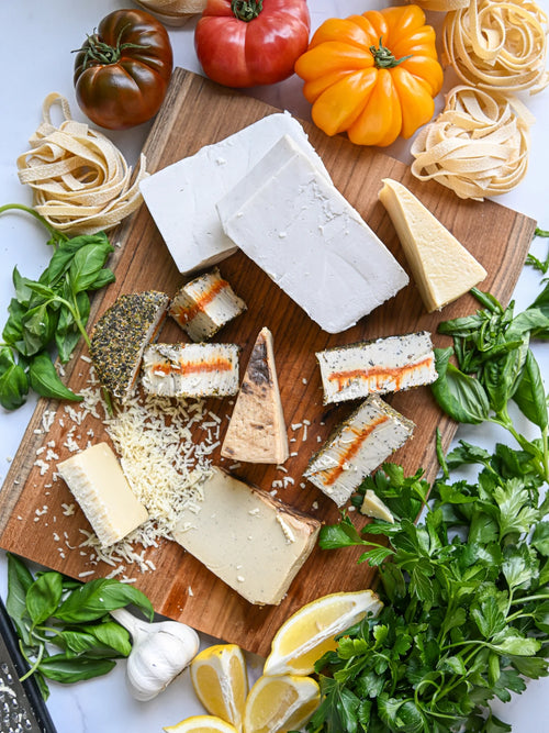 Assorted cheeses, pasta, and fresh herbs on a wooden board with lemons and tomatoes.