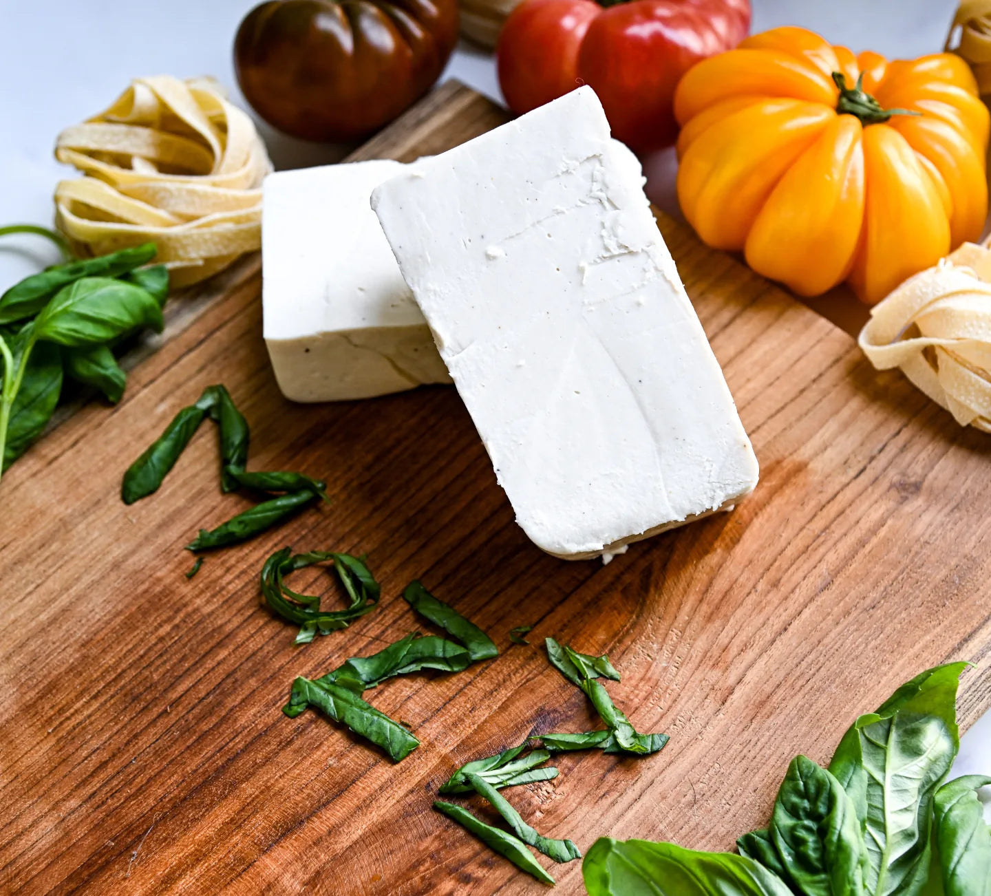 Block of Mozzarella on a wooden board with fresh basil leaves and colorful tomatoes.