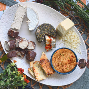 A cheese board displaying the 6 cheeses in our October Fromagier Box: Apple Cinnamon Chevre, Brie, Pimento, Pepper Jack, Tomato Herb Fromage, and Parmesan. Surrounded by flowers and fall-themed decorations.