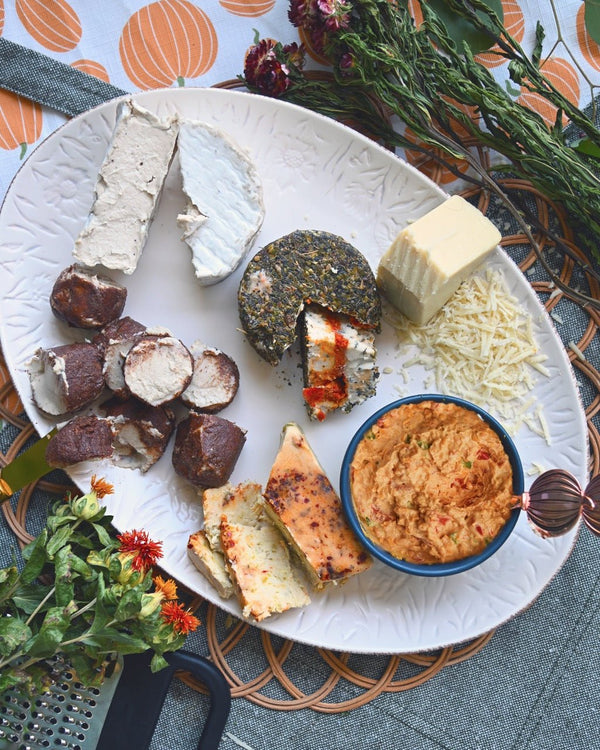 A cheese board displaying the 6 cheeses in our October Fromagier Box: Apple Cinnamon Chevre, Brie, Pimento, Pepper Jack, Tomato Herb Fromage, and Parmesan. Surrounded by flowers and fall-themed decorations.