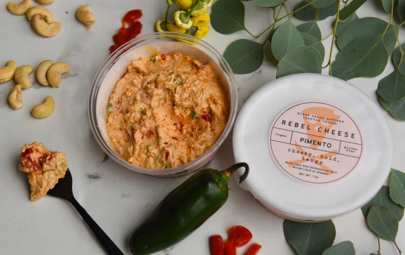 Container of Rebel Cheese Pimento on a marble surface with cashews and a jalapeño pepper.