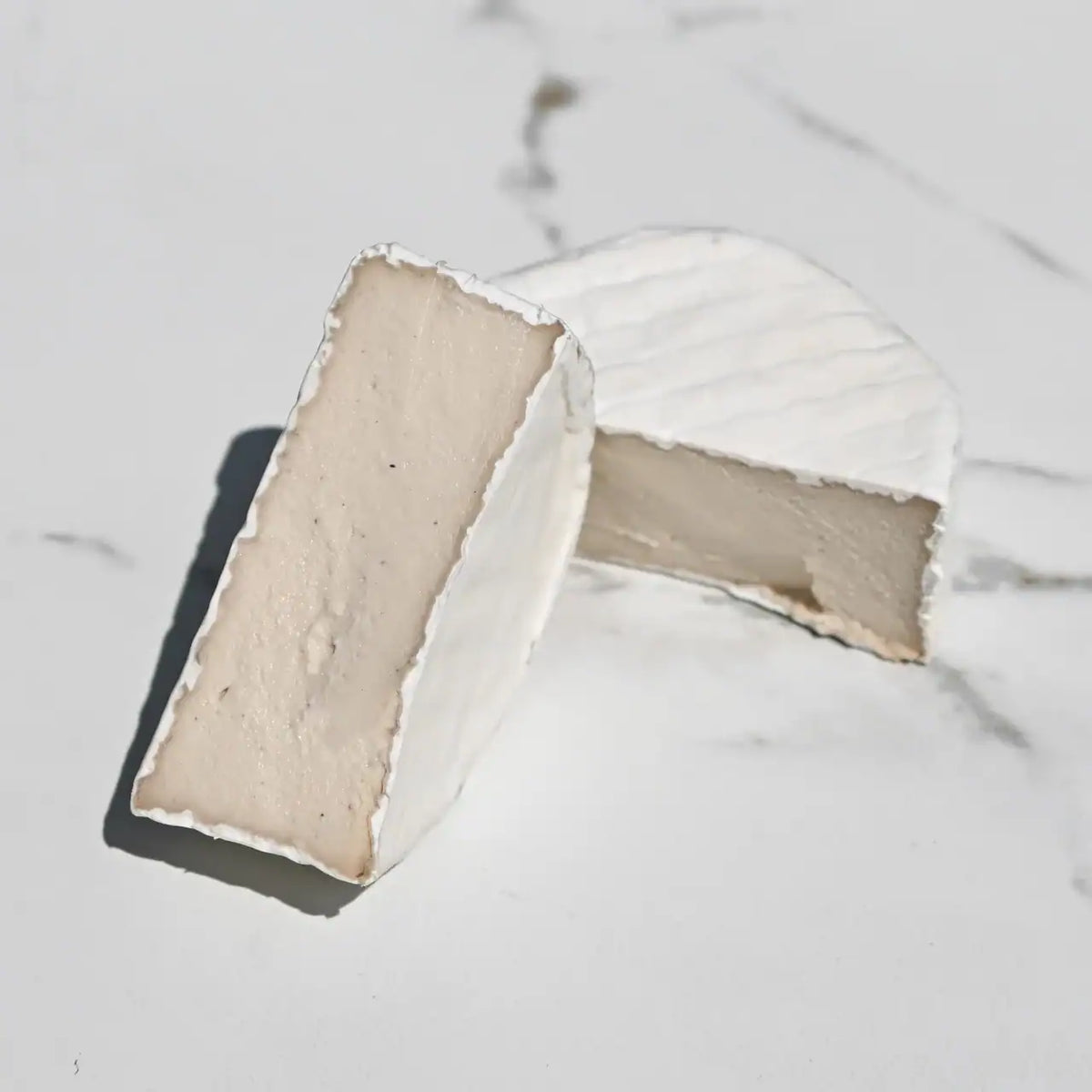 Two wedges of soft white Brie cheese on a light marble surface, with one wedge cut and standing upright to show the pale creamy interior and the other lying on its side.