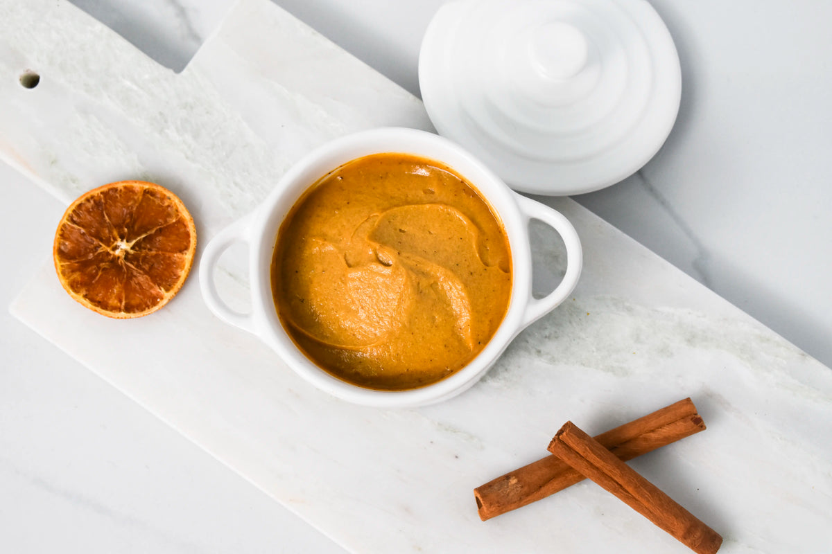Close-up of Rebel Cheese plant-based Pumpkin Spice Cream Cheese swirled in a white crock with cinnamon sticks and a dried orange slice on marble.