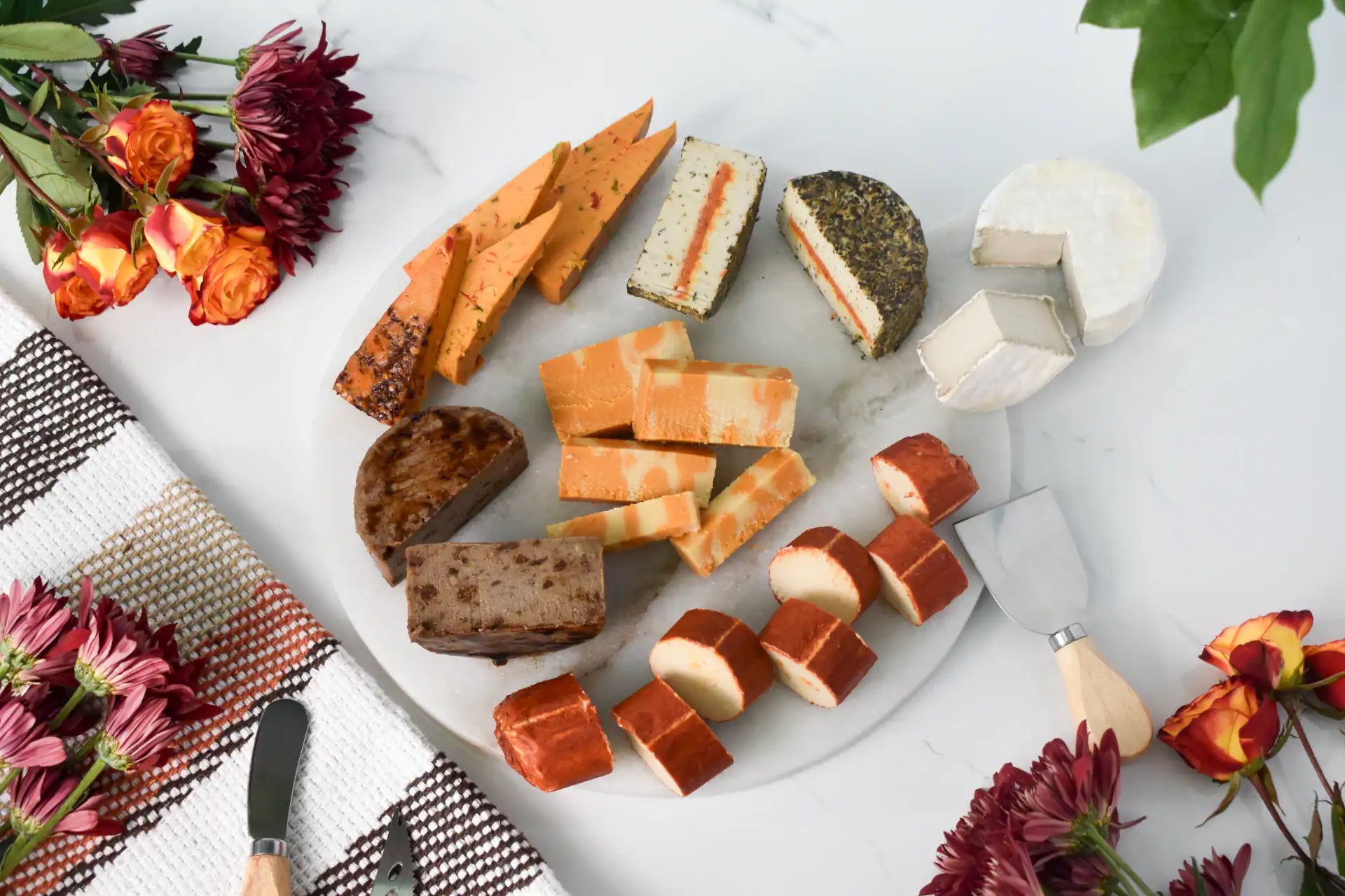 An overhead flat-lay of a charcuterie board with assorted cheeses on a white marble board, styled with flowers and a plaid napkin on a white background.