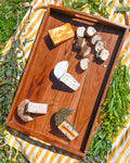 Wooden tray with various types of cheese on a striped outdoor tablecloth.