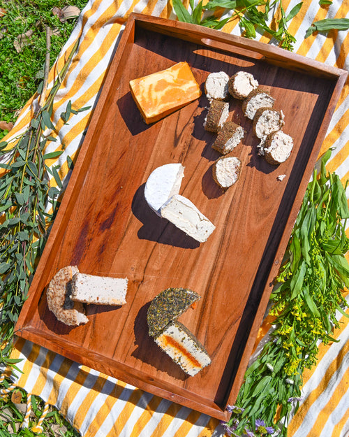 Wooden tray with various types of cheese on a striped outdoor tablecloth.