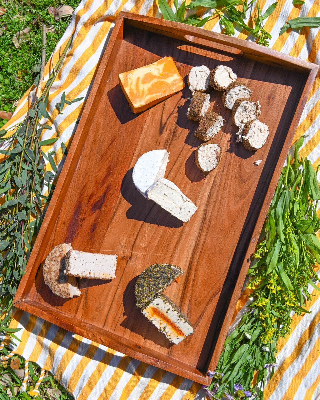 Wooden tray with various types of cheese on a striped outdoor tablecloth.