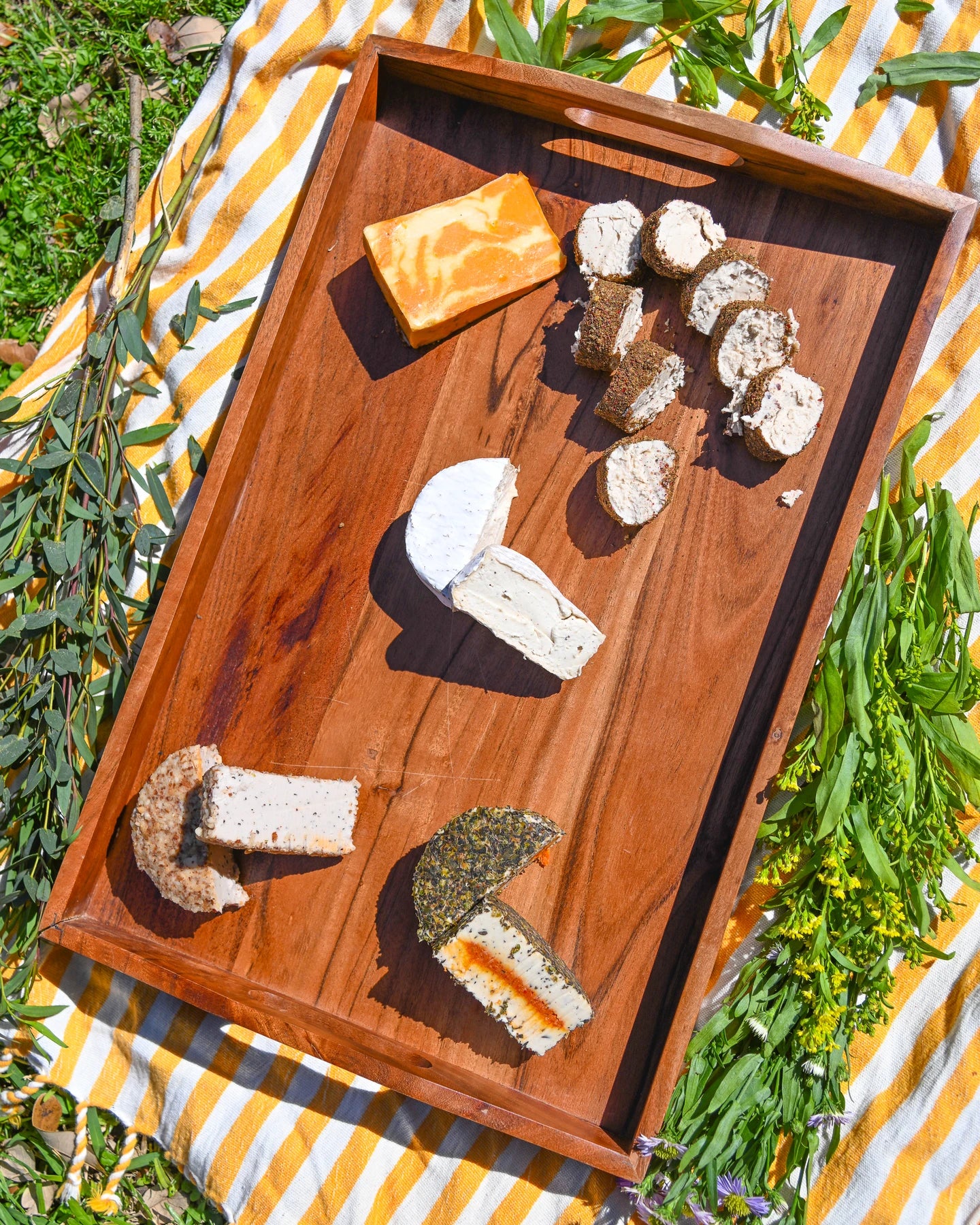 Wooden tray with various types of cheese on a striped outdoor tablecloth.