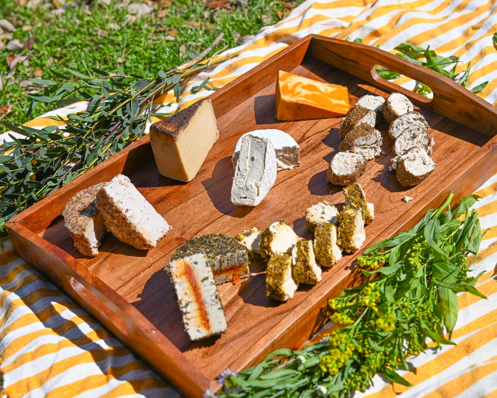 Wooden tray with assorted cheeses on a striped yellow and white blanket outdoors.