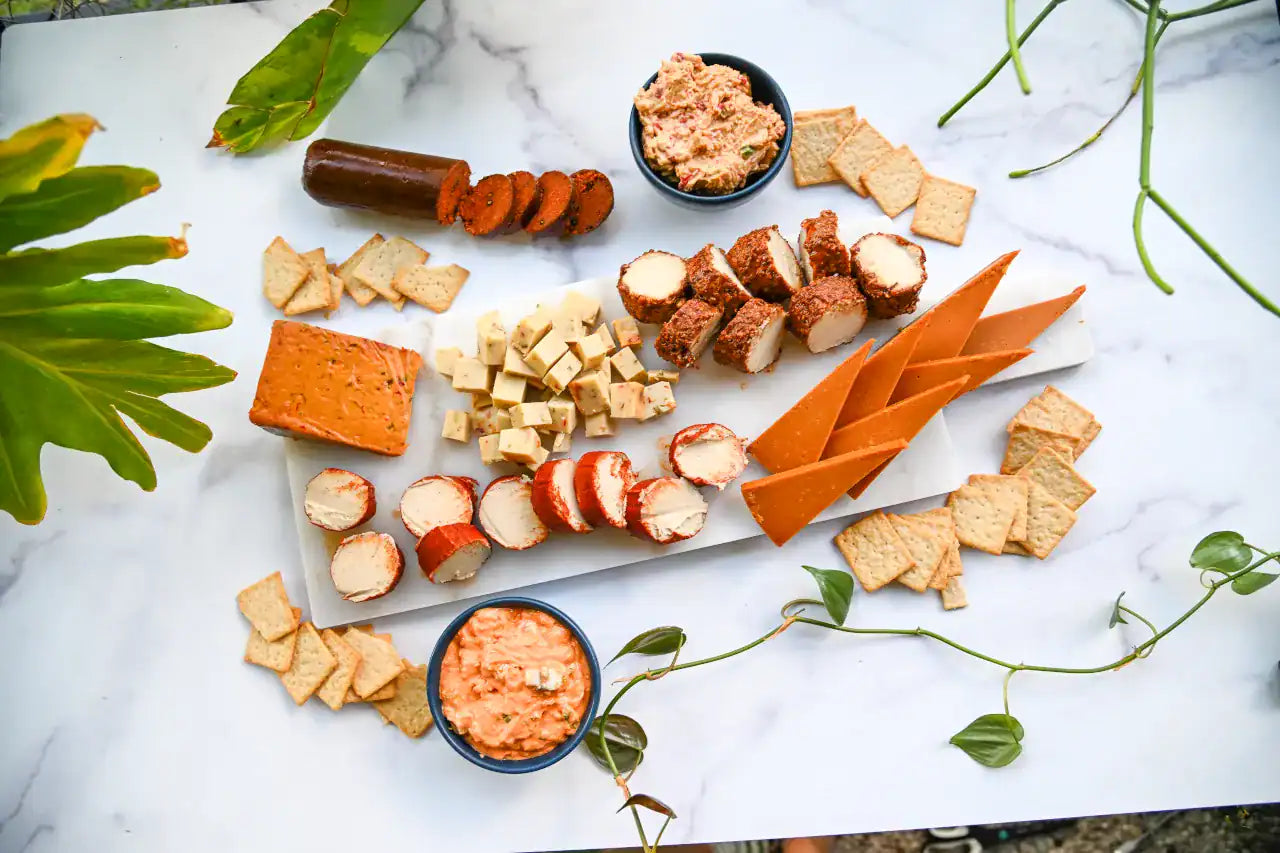 Assorted dairy-free cheese, dips, crackers, and vegan salami on a marble surface with green leaves.