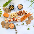 Assorted dairy-free cheese, dips, crackers, and olives on a marble surface with green leaves.