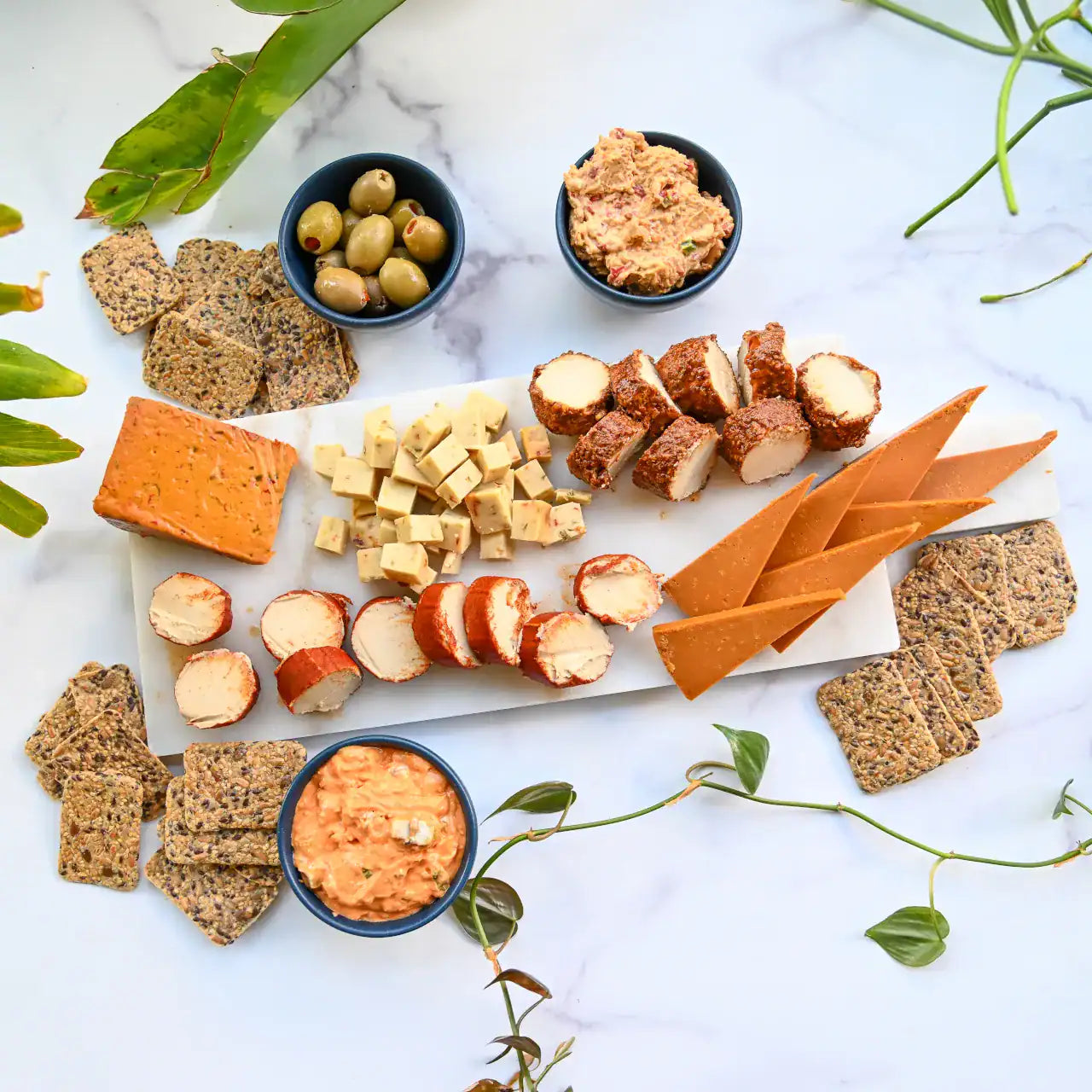 Assorted dairy-free cheese, dips, crackers, and olives on a marble surface with green leaves.
