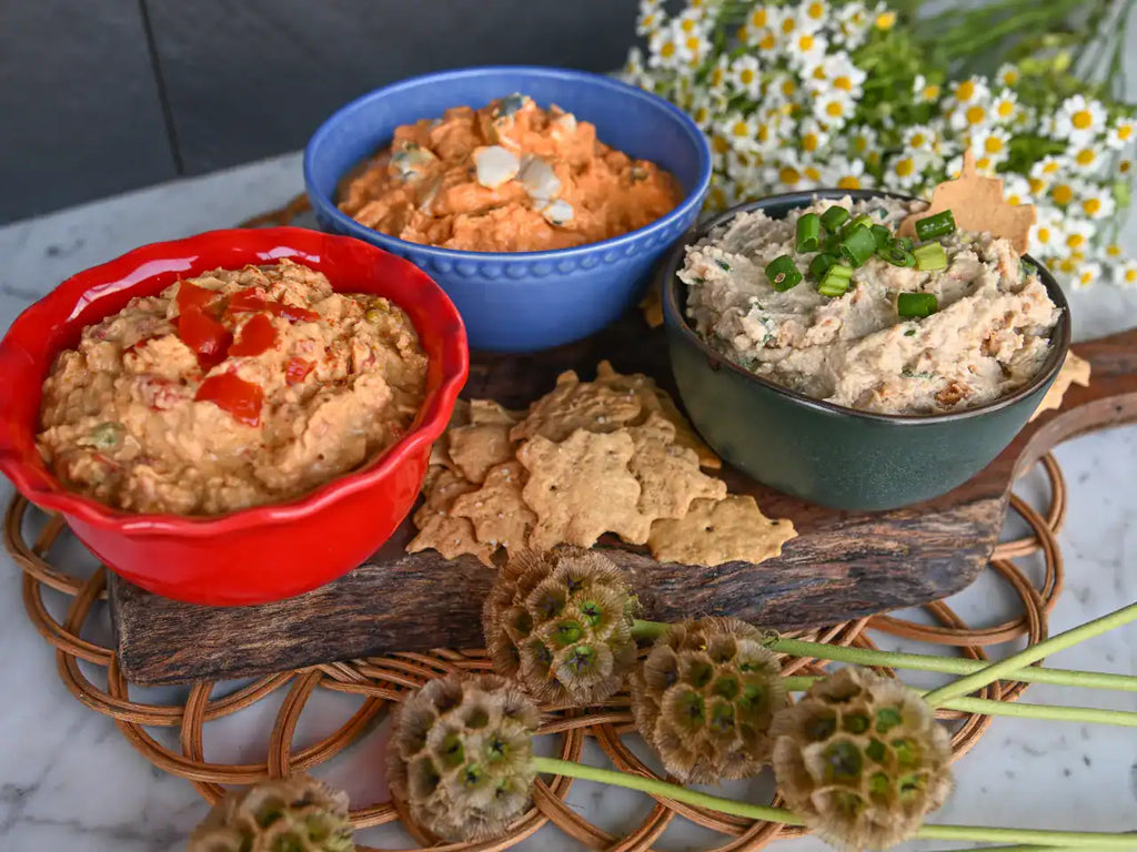 Three bowls of dip arranged on a wooden board with crackers, set on a woven tray with garlic flowers in the foreground and white daisies in the background.